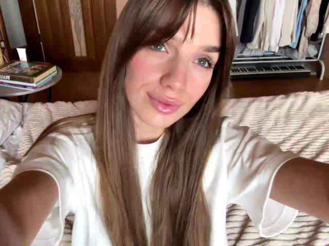 HildeLautner, com cabelos castanhos longos, olhos azuis, lábios rosados, vestindo uma camisa branca, deitada em uma cama, sorrindo para a câmera, com os braços estendidos, tirando uma selfie..