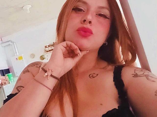 Pauleette Roux mit rotem Lippenstift, voller Lippen, einem schwarzen BH, Tätowierungen und langen braunen Haaren, die sich an ihr Kinn fasst, trägt Ohrringe im Stecker-Stil und ein Armband. Ihre Brüste sind sichtbar..
