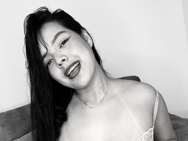ViolettaRouse with long dark hair, sticking out her tongue, smiling, wearing a lace bra, necklace, and having small, visible breasts in a black-and-white photo.