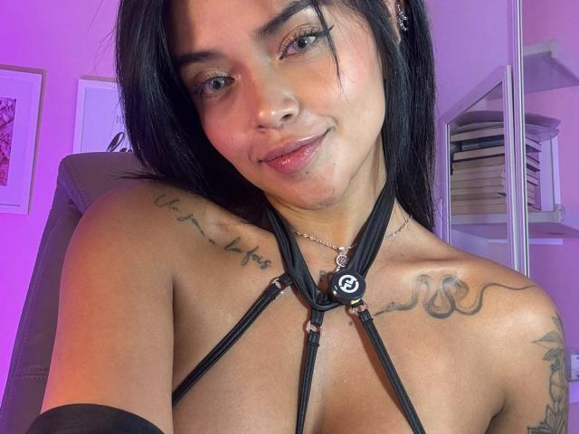 MajoJones, avec ses cheveux noirs, portant un harnais noir à bretelles, laissant apparaître des tatouages sur ses épaules, et un léger sourire, ses petites seins visibles, dans une pièce éclairée en rose..