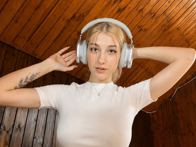 Georgann Whatcott, com cabelo loiro, vestindo uma camiseta branca, fones de ouvido prateados, braços levantados, tatuagem no antebraço esquerdo, olhando para a câmera, teto de madeira, painéis de madeira, colar, cabo visível..
