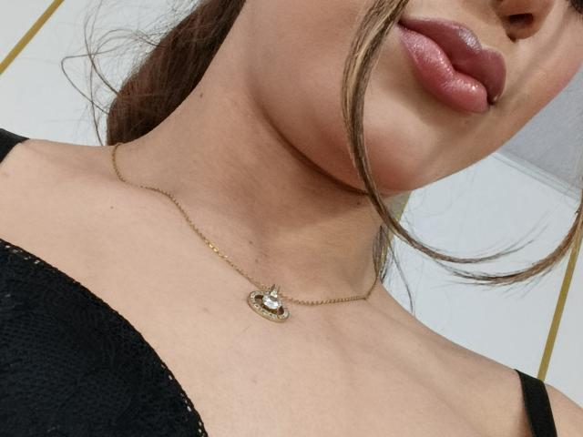 AliceCoolenk's face, showing glossy pink lips, a gold necklace with a small gem, and black lace bra. Her brown hair is partially tucked behind her ear.