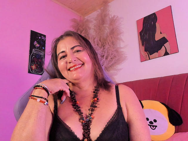 Paloma Lombardi, une femme d'âge moyen avec des cheveux bruns, souriante, portant un soutien-gorge en dentelle noire, un collier de perles rouges, des bracelets rouges, assise sur une chaise rouge, avec un coussin jaune orné d'oreilles noires, devant un mur rose..