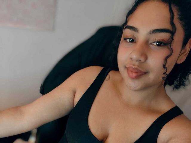 VickyCassel with curly black hair, brown eyes, and tan skin, wearing a black tank top, showing her small tits, pouty lips, and a subtle smile, in a selfie.