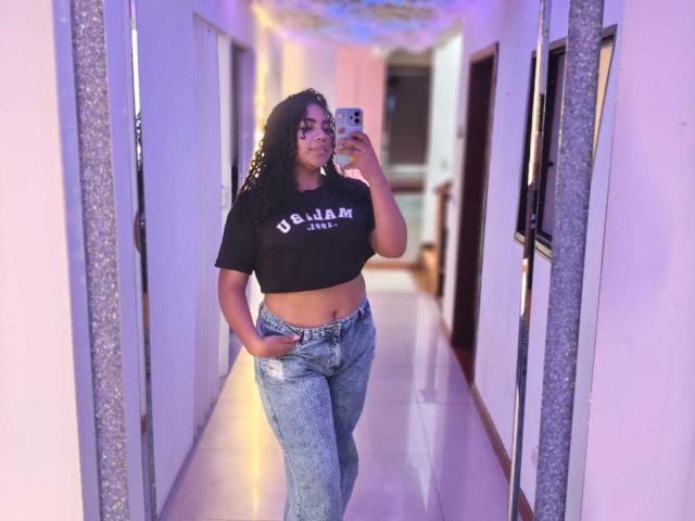 VickyCassel with curly hair, wearing a black crop top and blue jeans, standing in a hallway, holding a phone, hands in pockets, smiling, showing a bit of her ass.
