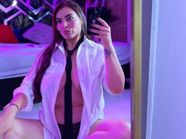 MarianaFoxyHot, with long brown hair, sitting, wearing an open white shirt, black tie, and black panties, showing her tits, holding a phone, in a purple-lit room.