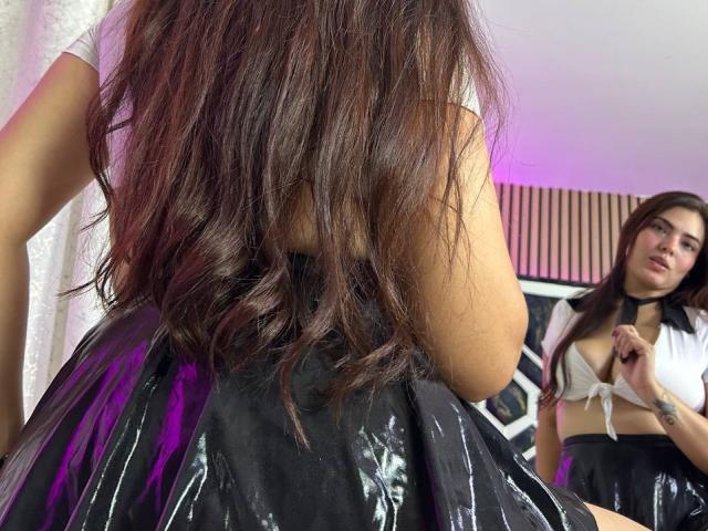 MarianaFoxyHot with long brown wavy hair, wearing a black shiny skirt and white tied top, standing in front of a mirror, showing her bare ass and small tits.