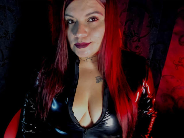 MoonDark69 with red hair, black latex top, deep cleavage, tattoo on chest, dark lipstick, smiling, choker necklace, red and black lighting, glossy texture, sexy, confident.