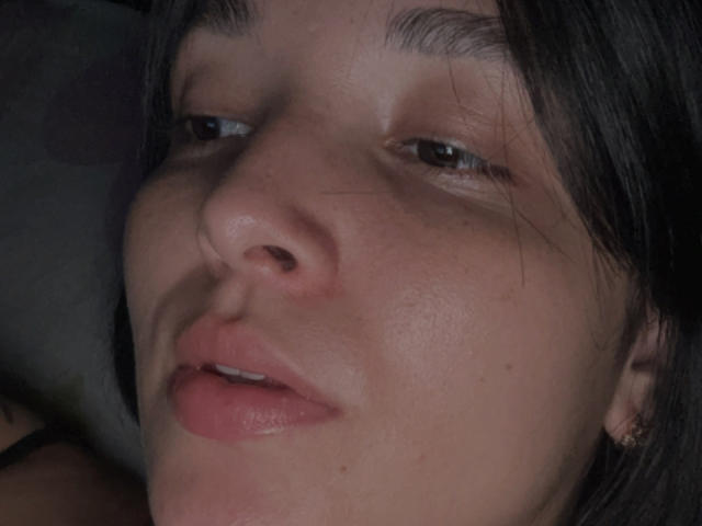 SarahGood's face with slightly parted pink lips, brown eyes, and dark brown hair, showing a subtle nose and small ear with a gold hoop. Her skin is light with freckles.