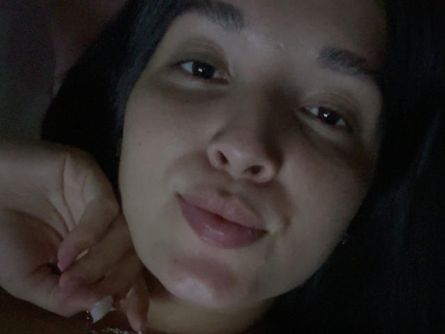 SarahGood's face, showing dark hair, brown eyes, and slightly parted pink lips, with her hand touching her neck, in a dimly lit, close-up shot.