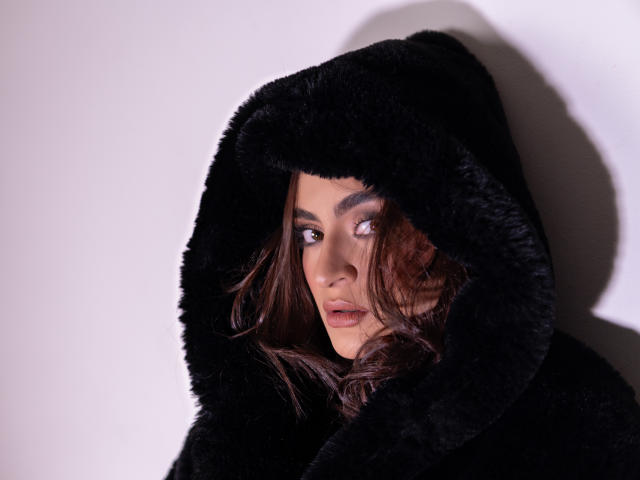 AruaMiller with wavy brown hair, wearing a black fluffy hood, looking to the side with a sultry expression, soft light highlighting her face.