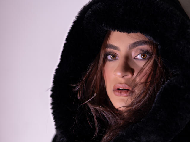 AruaMiller with brown eyes, wearing a black furry hood, her lips slightly parted, brown hair peeking out, looking directly at the camera with a sultry expression.