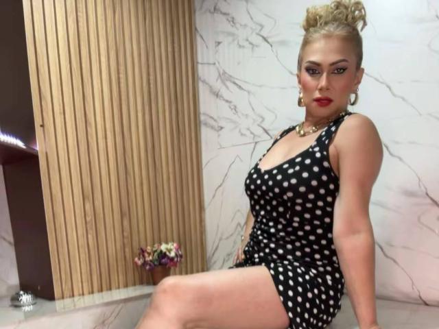 MisseScarlet com o cabelo loiro preso, batom vermelho e brincos de argola, vestindo um vestido preto com bolinhas, sentada com as pernas cruzadas, mostrando o bumbum. Parede de mármore e painel de madeira ao fundo..