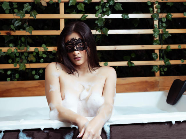 AruaMiller with dark hair, wearing a black lace mask, sits in a bubbling hot tub, her soapy tits partially visible, hands resting on the tub's edge.