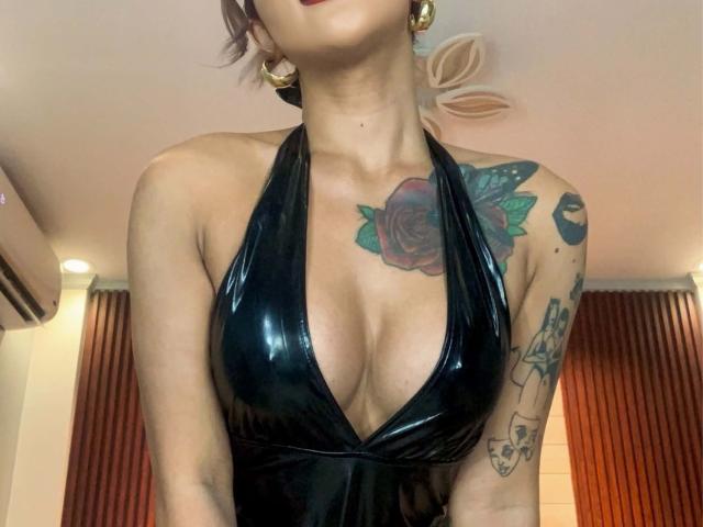 SeductiveSophia with shiny black halter top, large tits, red rose tattoo on chest, green leaves, gold earrings, and arm tattoos, head tilted back, red lips.