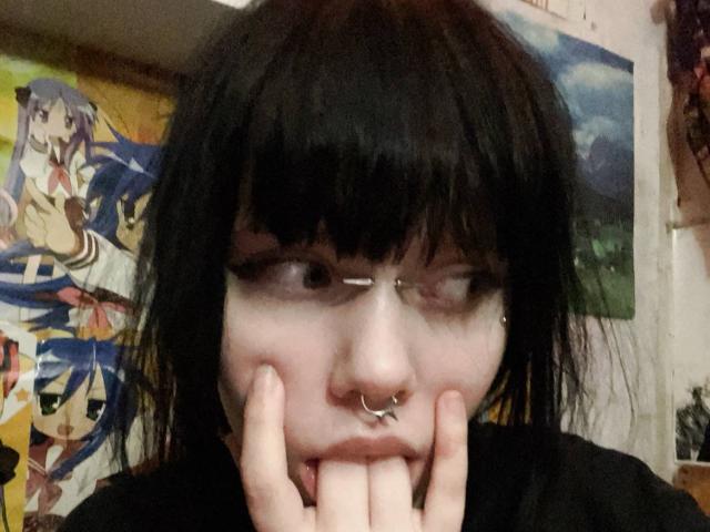 MariannaWarren with black hair, nose ring, and septum piercing, biting her finger, wearing black eyeliner, in a bedroom with anime posters. Her hand covers her mouth slightly.