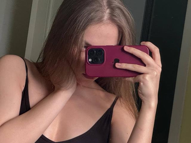 KasieRoshia with long brown hair, black tank top, holding a pink phone covering her face, showing her small tits, taking a mirror selfie, in a dimly lit room.