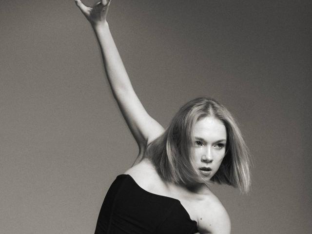 Black-and-white photo of KasieRoshia with shoulder-length blonde hair, raising her right arm, wearing an off-shoulder black top, showing her bare shoulder and upper chest.