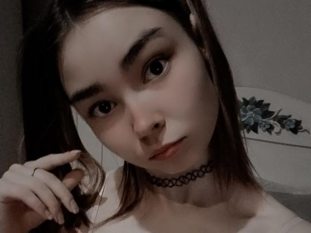 LelaVivo with pale skin, dark eyebrows, and brown eyes, wearing a black choker and white top, holding brown hair, showing a subtle, serious expression.