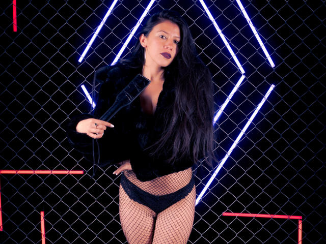 SandyQuinnX with long black hair, wearing a black fur jacket, fishnet stockings, and black lace panties, holding a microphone, showing her tits, against a neon geometric pattern.