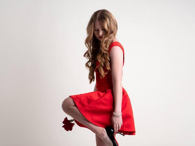 AkilahGhil in a red dress, holding a red rose, bends one knee, showing her slim legs and long wavy blonde hair, wearing black heels, white bracelet.