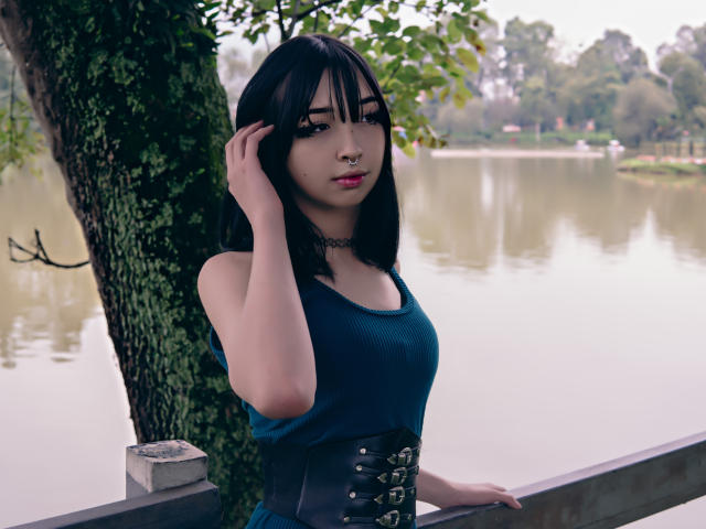 Vickmon with black hair, blue top, black choker, and corset, standing by a tree, hand in hair, looking at camera, nose ring, sexy, water background, pale skin.