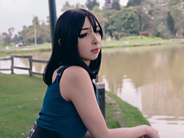 Vickmon has black hair, wearing a blue tank top, with visible small moles on her face, sitting by a pond, looking over her shoulder, hands on a wooden railing.