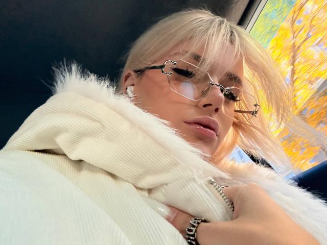 VedaVasi with blonde hair, glasses, and a fur coat, showing a fluffy white sweater, looking slightly down with a subtle smile, sunlight through the window.