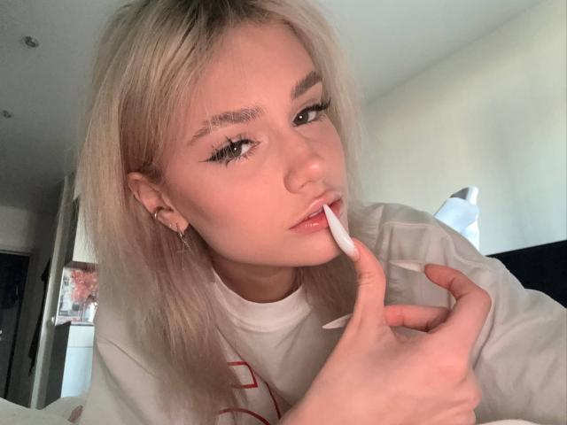 VedaVasi with blonde hair, applying white lipstick to her slightly parted pink lips, looking at the camera, wearing a white shirt, in a softly lit room.