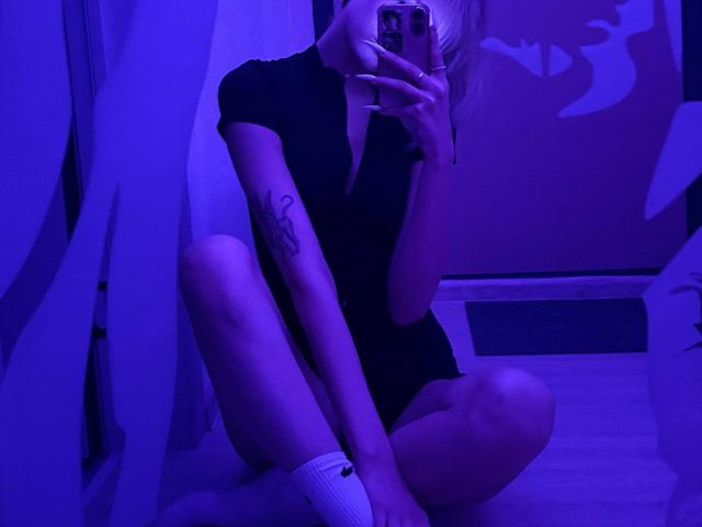 VedaVasi sits on the floor in a black dress, holding a phone, showing a tattoo on her arm, wearing white socks, in blue-lit room, covering her tits with her hand.