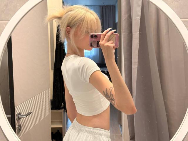 VedaVasi with blonde hair in a ponytail, white crop top, and gray pants, showing a tattoo on her right arm, taking a mirror selfie with a pink phone.
