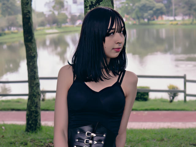 Vickmon with black hair, black tank top, and leather corset, standing in a park, has small tits, nose ring, and a serious expression, looking to the side.