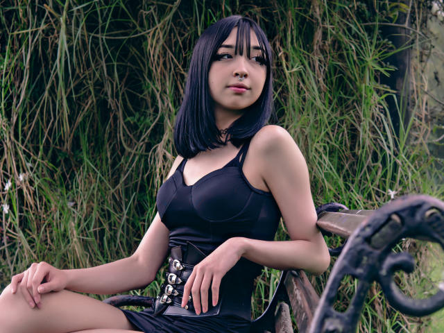 Vickmon with black bob, wearing a tight black dress and corset, sitting on a metal bench, looking up with a slight smile, green grass and plants in the background.
