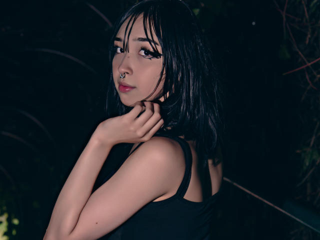 Vickmon with dark wet hair, nose ring, and black tank top, looking back over shoulder, hands near neck, soft lighting, dark background. Boobs not visible, focus on face and upper body.