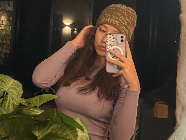 JuliethCourtouis with wavy brown hair, wearing a brown knit beanie and purple long-sleeve top, holding a phone with a leaf design, in a dimly lit room.