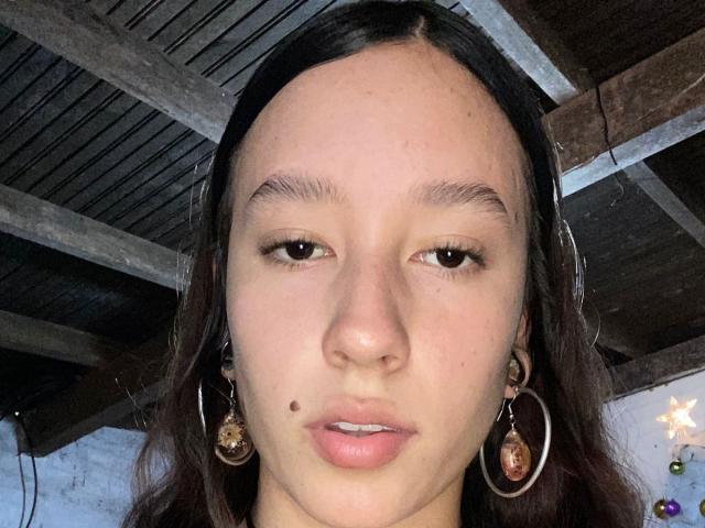JuliethCourtouis has a small mole on her left cheek, brown eyes, and long dark hair. She's wearing large hoop earrings with ornate pendants. Her face is centered.