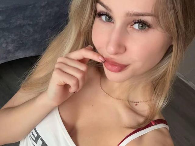 MilagrosEdmisten has blonde hair, green eyes, and is touching her pink lips with her finger. She's wearing a white top with red straps, and a thin gold necklace.