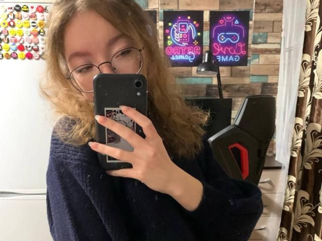 ClaireSpisak with curly blonde hair, glasses, and a navy sweater, taking a mirror selfie with her phone, showing her nose and mouth. Gaming chair and neon signs in background.