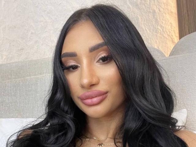 LunessaNoir tiene el cabello oscuro y ondulado, cejas pobladas, lápiz labial rosa y pestañas largas, y mira a la cámara con confianza, con una ligera sonrisa. Su piel es morena y tersa..