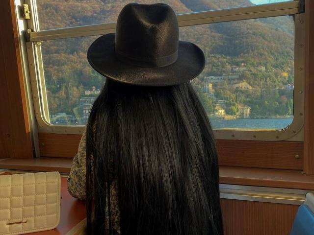 LunessaNoir, com longos cabelos pretos, usando um chapéu de cowboy preto, sentada em um trem, de frente para uma janela, mostrando uma vista de montanhas e florestas lá fora..