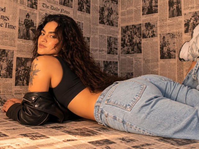 UnfulFilled with curly brown hair, black crop top, and blue jeans, lying on newspaper, black leather jacket off, showing a tattoo on her right shoulder, looking over her shoulder.