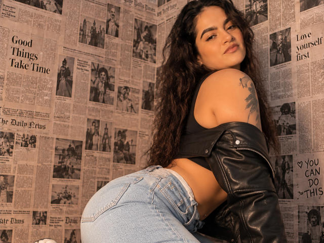 UnfulFilled with long curly brown hair, black crop top, and blue jeans, leans forward showing her ass, wearing a black leather jacket, tattoo on her right shoulder.