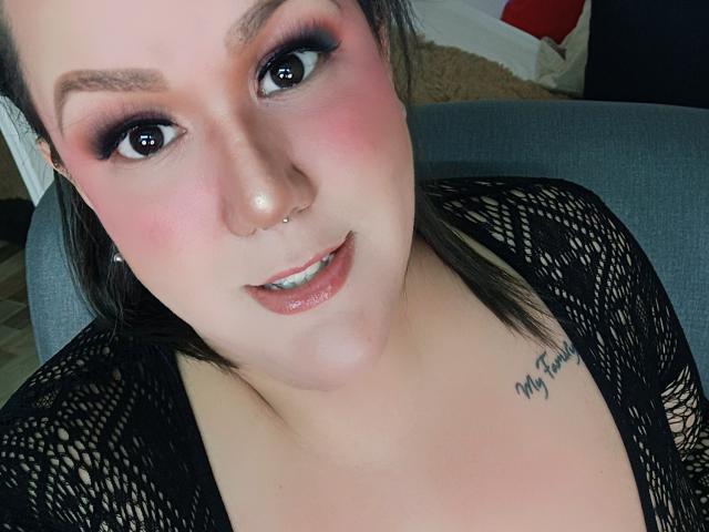 GiiannaMorrys mit rosigem Rouge, dunklem Eyeliner und glänzendem Lipgloss, trägt eine schwarze Spitzenbluse, einen Nasenring und ein Tattoo mit der Aufschrift "My Forever" auf ihrer rechten Brust..