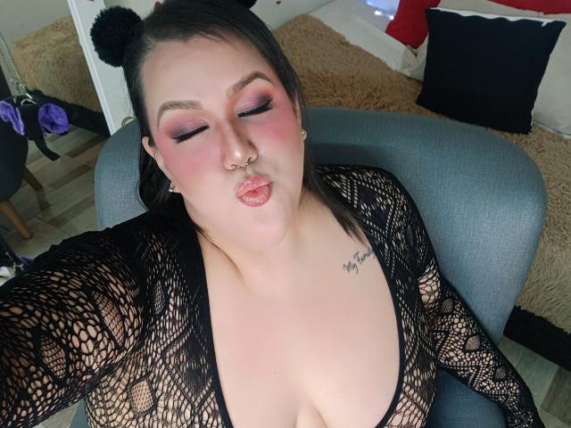 GiiannaMorrys with puffy pink lipstick, black fishnet top, and nose ring, pouting lips, black hair with bear ears, tattoos, and large boobs, sitting in a gray chair.