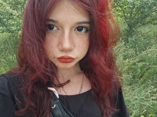 MarioChoudhary has red hair, freckles, and orange lipstick, wearing a black top with a silver necklace, looking serious with a slightly parted lip and a black bag strap.