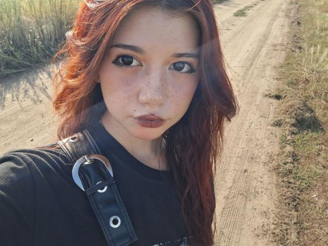 MarioChoudhary with red hair, freckles, and dark lipstick, wearing a black shirt and leather strap, looking directly at the camera, standing on a dirt path.