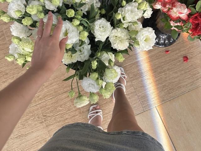 Le mani di Diantrisy che toccano un bouquet di ortensie bianche e verdi, indossa sandali bianchi con cintini, una gonna di jeans grigia, pavimento in legno, un anello alla mano sinistra, riflesso sul pavimento, fiori rossi sullo sfondo..