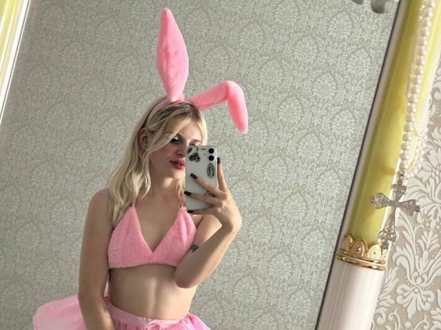 FallenAngelia with blonde hair, pink fluffy bunny ears, pink bra, and pink skirt, taking a mirror selfie, showing her tits, holding a phone with a white case.