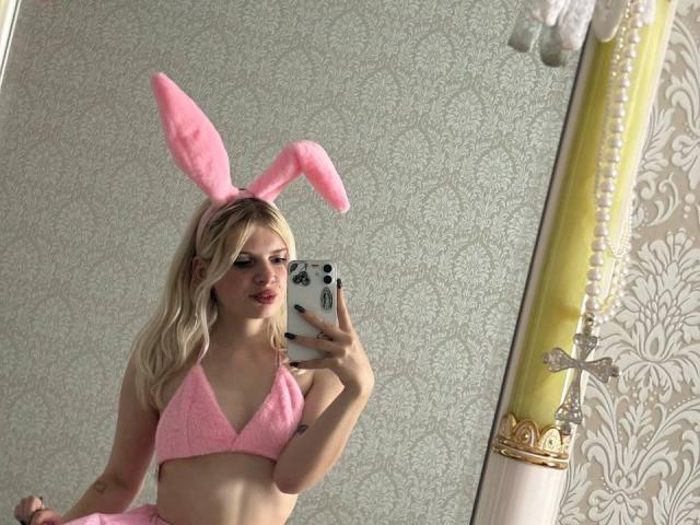 FallenAngelia with pink bunny ears, pink bikini top, blonde hair, holding phone, mirror selfie, showing small tits, pale skin, pink lips, white phone case, elegant room.