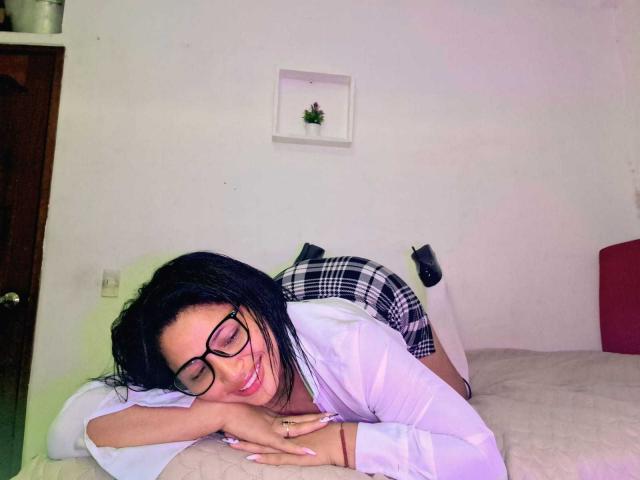 Roxane, une femme avec des cheveux noirs, des lunettes, une chemise blanche et une jupe à carreaux, allongée sur un lit, souriante, les fesses en l'air, portant des chaussettes blanches, la main droite sur l'oreiller, la main gauche sur le lit..